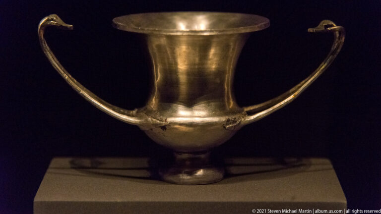 Polished silver drinking cup on a short foot with a flared rim and two high looping side handles.