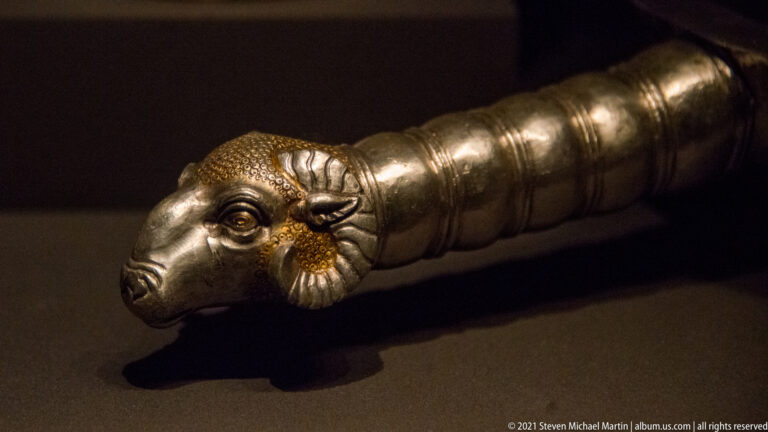 Close-up of a silver vessel handle ending in a detailed ram's head with curled horns, textured fleece, and ridged shaft.