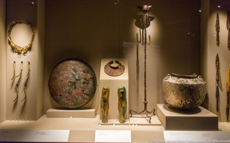 Grave goods from the Prince’s Tomb including a round shield, paired greaves, tripod, cauldron, wreath, and weapons.