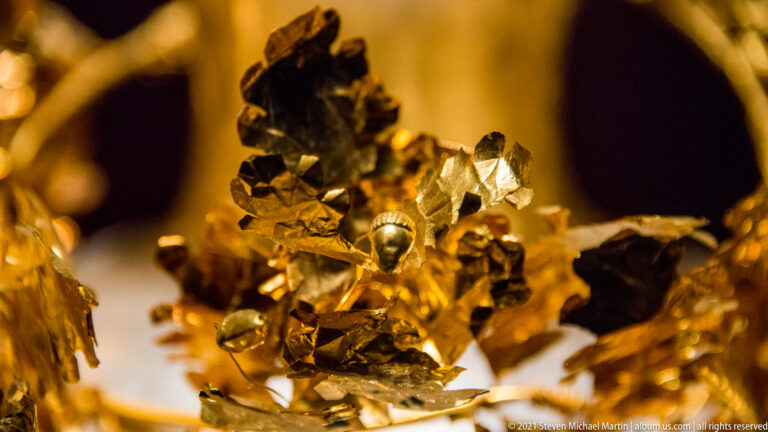 Close view of a gold wreath with crumpled leaves, small buds, wire stems, and overlapping botanical ornaments.