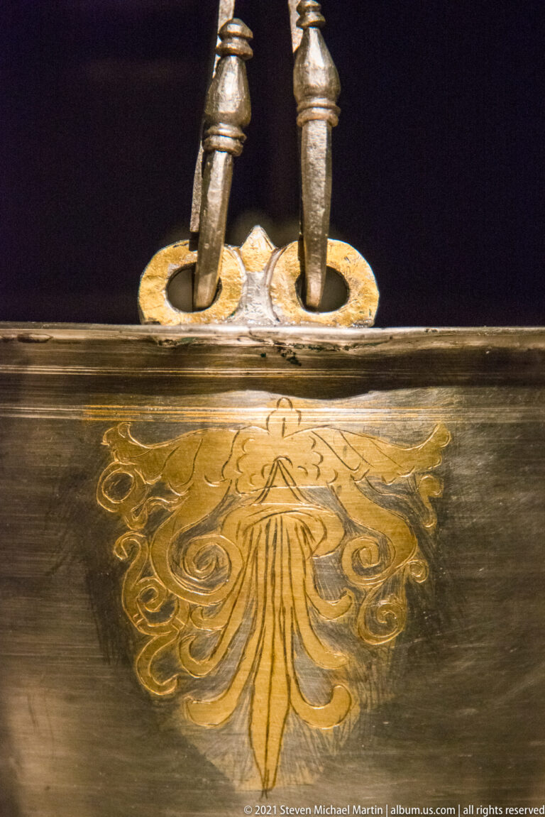 Close detail of a metal object with a gilded palmette motif below two vertical fittings and a shaped mounting plate.