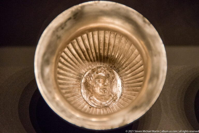 Shallow silver bowl viewed from above with radial fluting and a small human face medallion at the center.