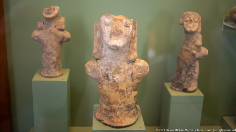 Three weathered terracotta human-like figurines displayed on separate stands, with the broad central figure closest to the camera.