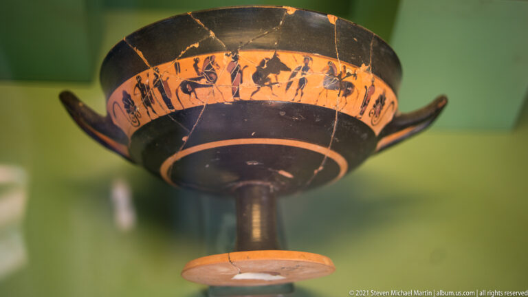 Black-figure kylix viewed from below, with a stemmed foot, two handles, and a continuous human-figure frieze.