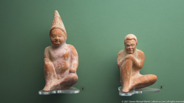 Two small seated terracotta figures, one wearing a tall pointed headdress, displayed side by side on clear mounts.