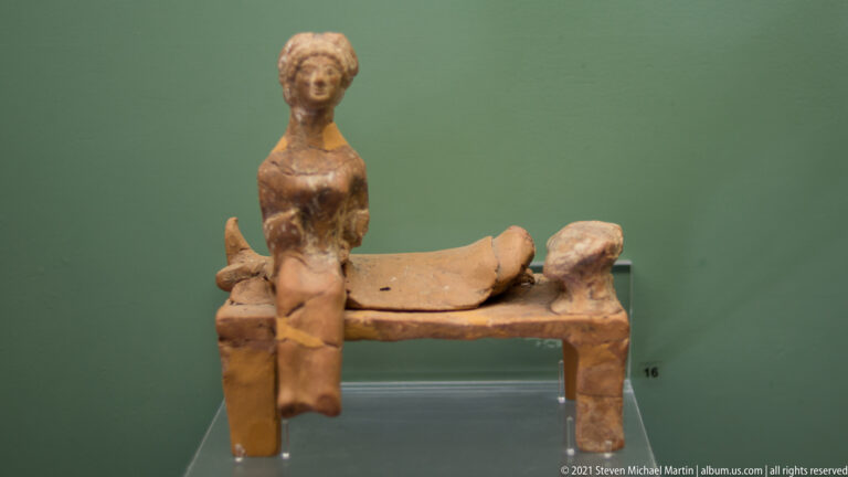 Terracotta female figure seated beside a low couch or bed-like platform with a smaller modeled form.