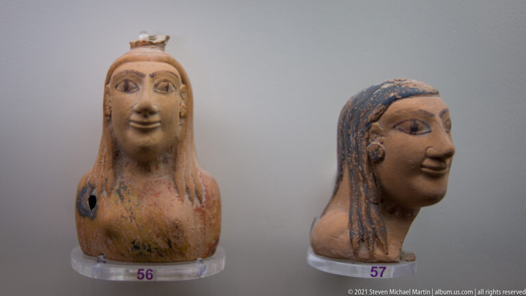 Two terracotta female head fragments with stylized faces and surviving dark-blue pigment in the hair.