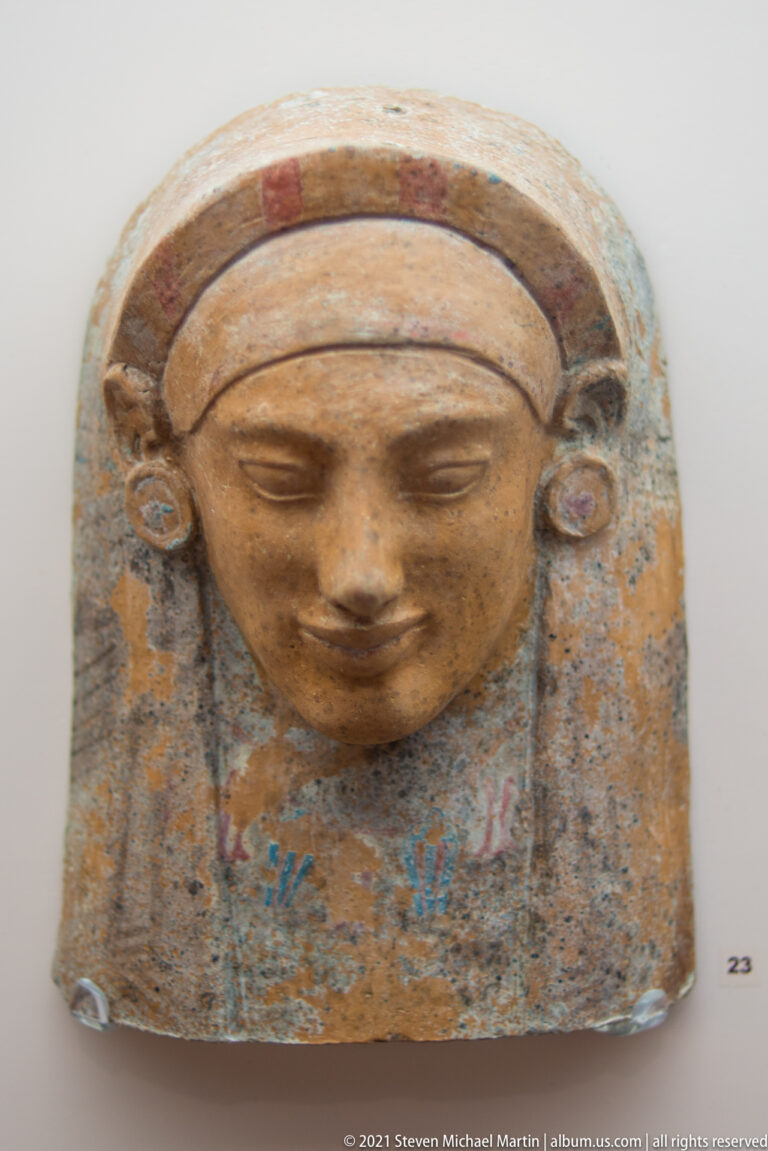 Rectangular painted terracotta plaque with a relief female face, curved headdress, and traces of blue and red decoration.