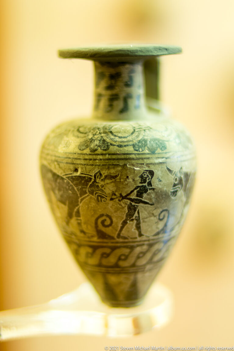 Small round ancient ceramic vessel with dark figure decoration, including a horse-like motif, beneath a broad flat rim.