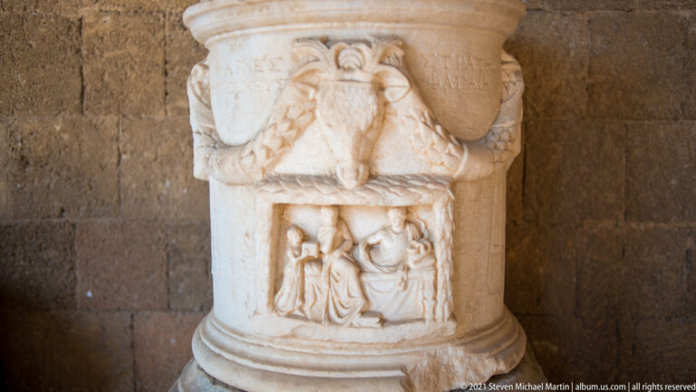 Cylindrical marble monument with inscriptions, an upper winged motif, and a lower relief panel containing human figures.