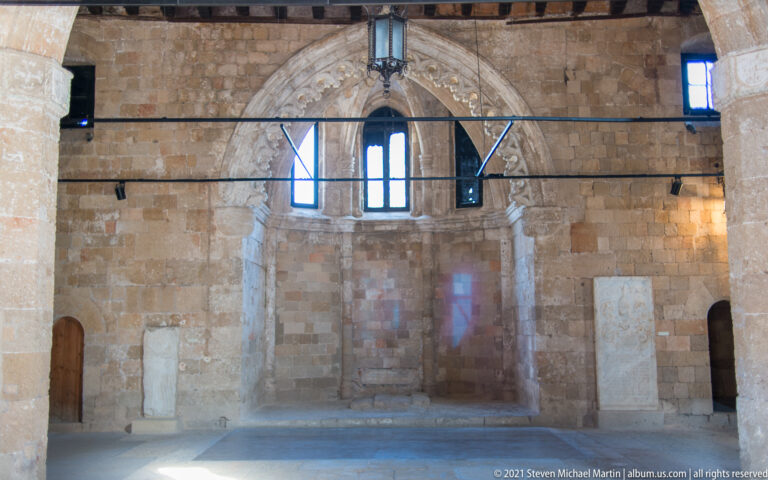 Large medieval stone hall with a pointed decorative arch, tall windows, timber ceiling elements, and sparse museum displays.
