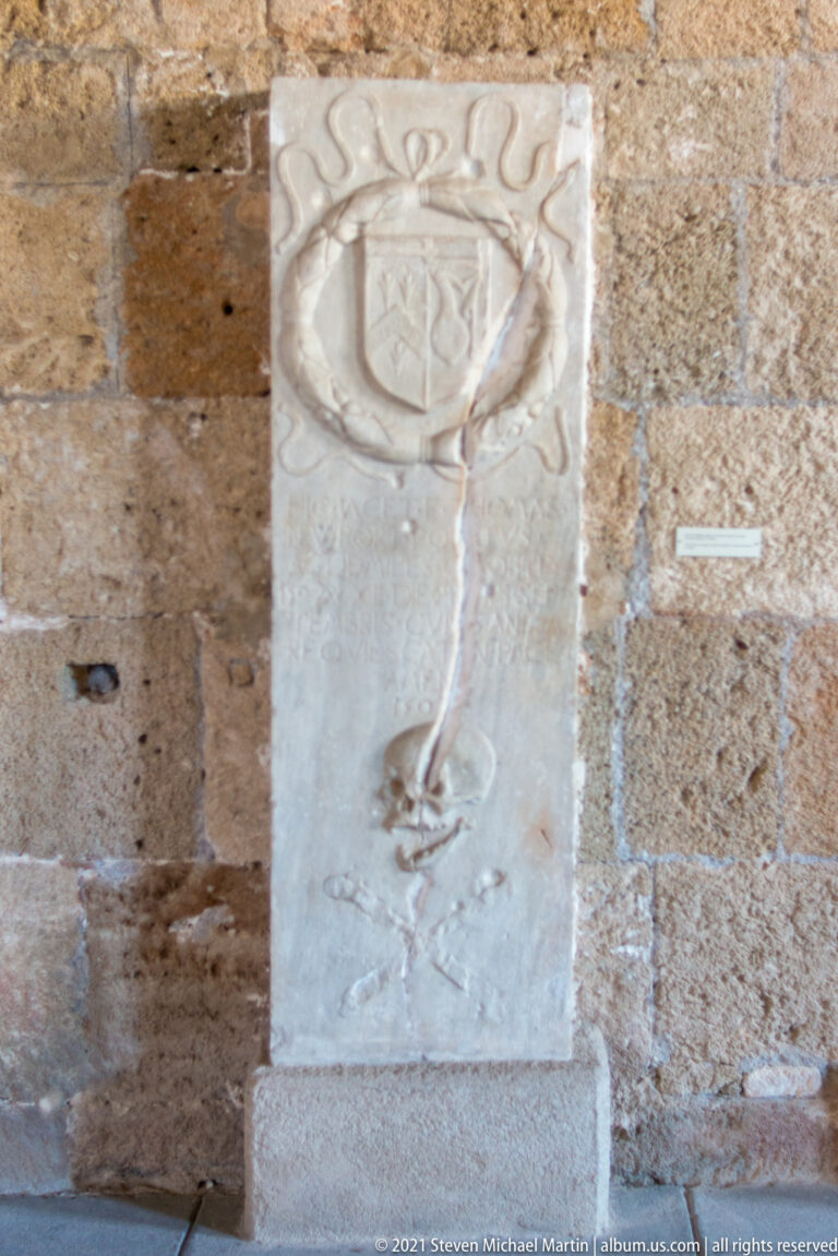 Tall pale stone slab carved with a shield inside a wreath and a long pendant ornament below.