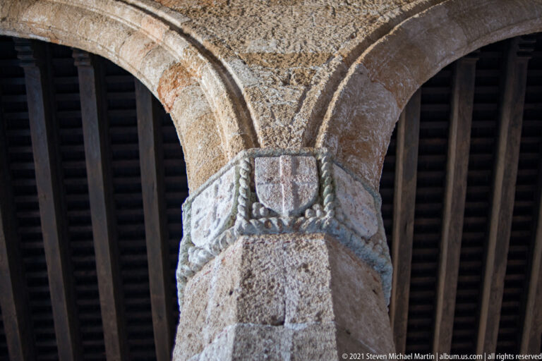 Carved stone capital with a central shield and foliage beneath intersecting arches and a dark timber roof.