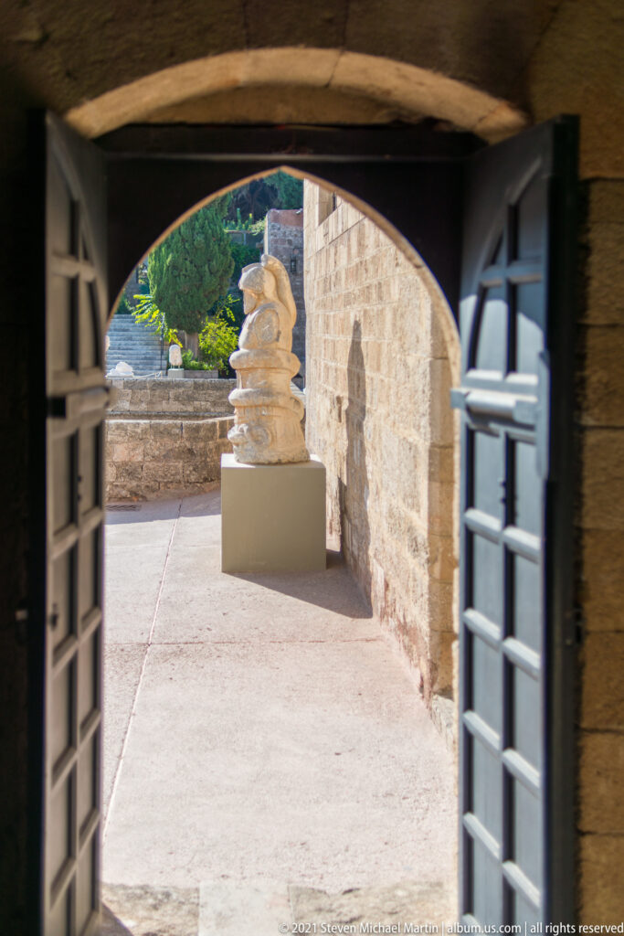 Dark arched doorway framing a sunlit stone passage and a stacked sculpture on a pedestal outside.