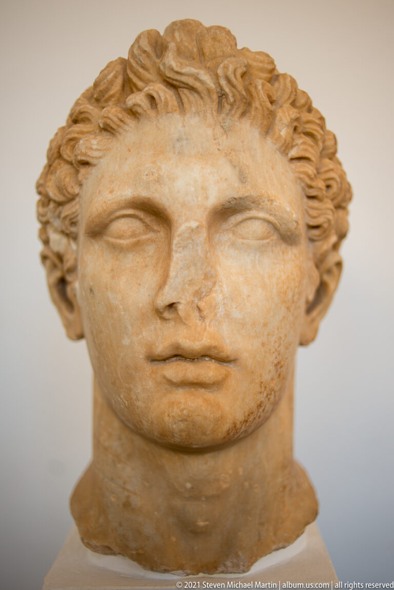 Frontal marble head of a youthful male with short curly hair, smooth cheeks, and an idealized expression.