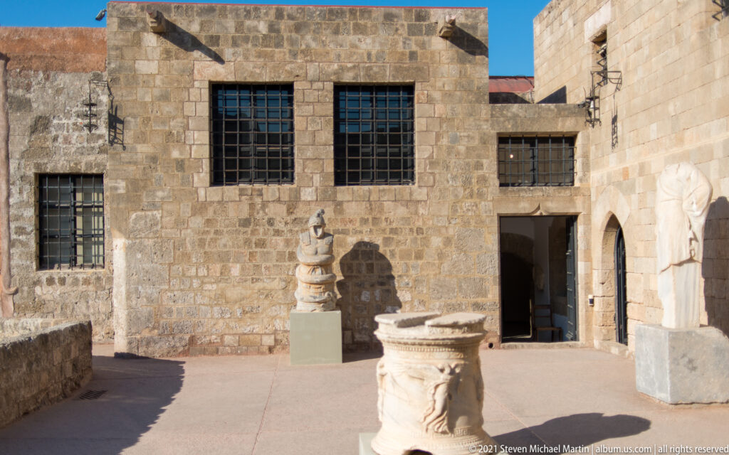 Sunlit stone courtyard containing several carved stone objects and sculptures on modern display plinths.