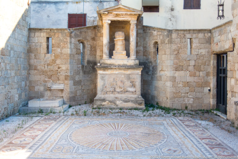 Pedimented stone monument against a courtyard wall above a mosaic pavement with a large radiating circular design.