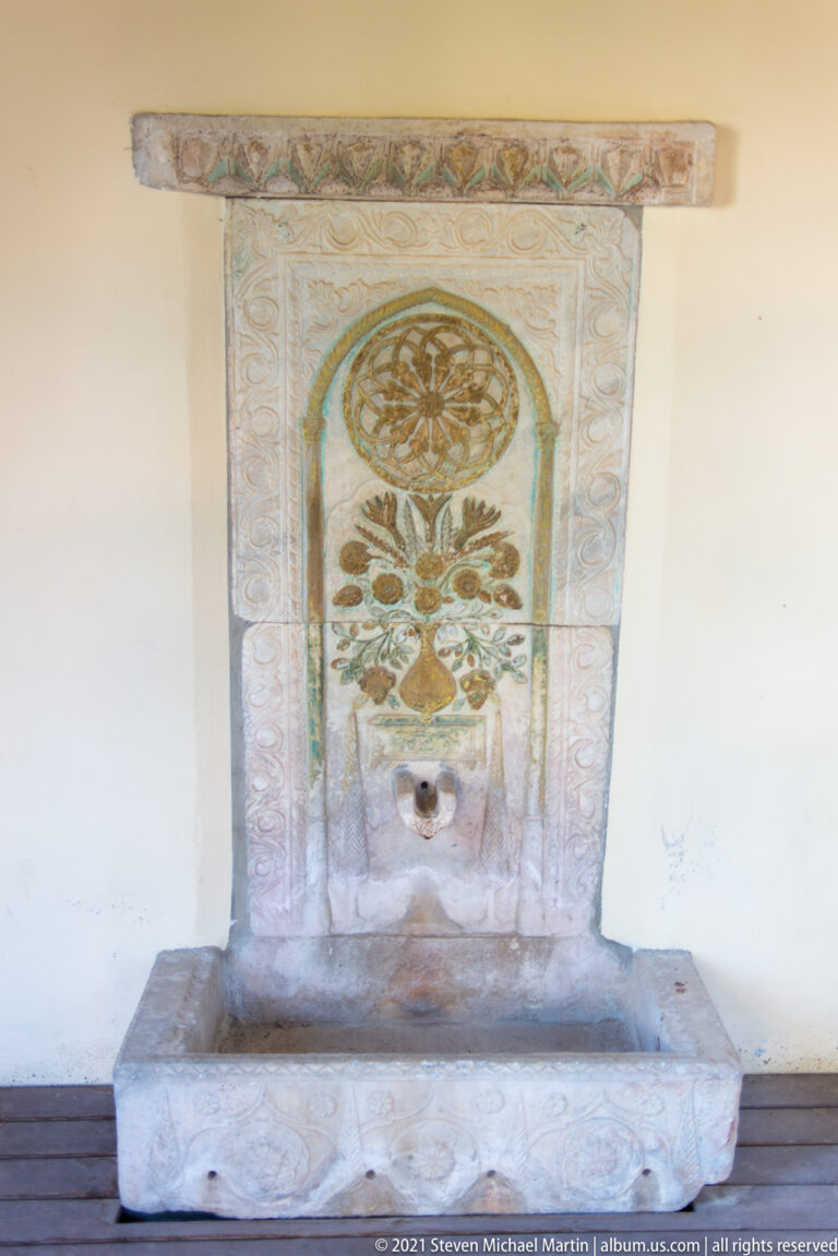 Stone wall fountain with rectangular basin, floral decoration, circular rosette, and a small water outlet.