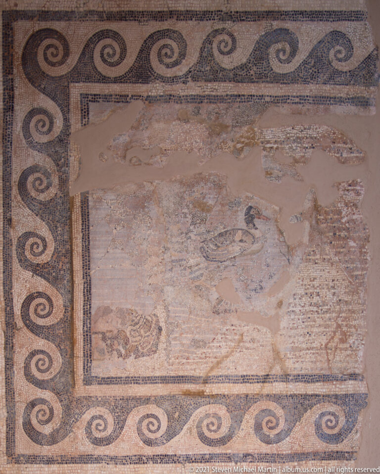 Fragmentary floor mosaic with a broad scrolling-wave border and worn remains of a central figural scene.