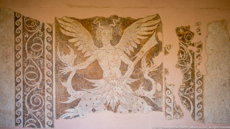 Fragmentary mosaic of a frontal winged female figure with spread wings beside ornate scrolling borders.
