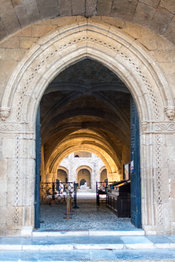 Carved pointed-arch doorway opening into a long vaulted stone hall with another bright arch in the distance.