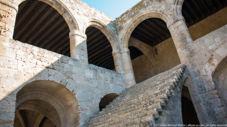 Upward view of a broad stone staircase leading to an upper gallery with tall arches and timber roofing.