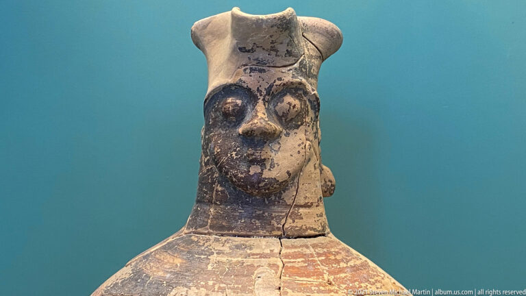 Close view of a painted ceramic vessel modeled with a human face beneath a pale flaring rim.