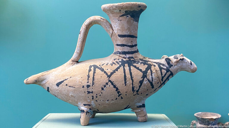 Bird-shaped ceramic vessel on small feet with tall neck, loop handle, modeled head, and dark painted lines.