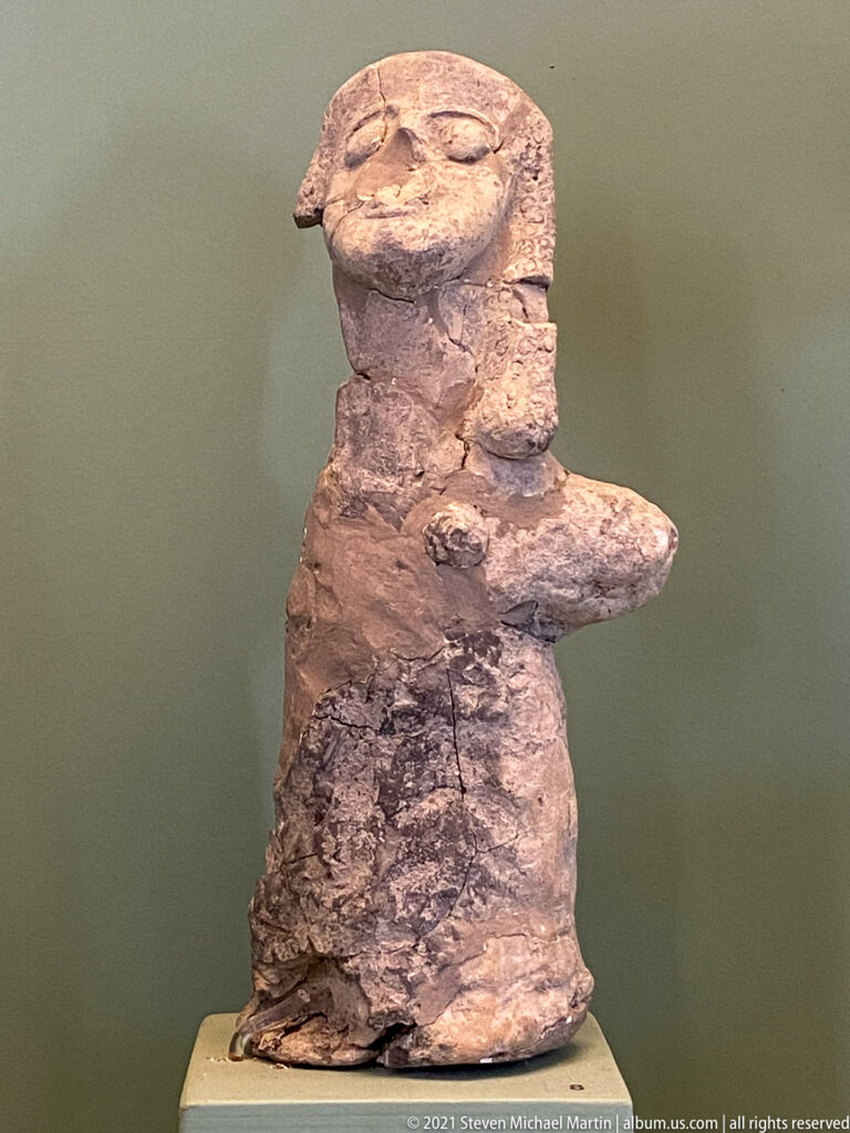 Weathered terracotta human figurine with broad head, projecting arms, and heavily eroded surface details.