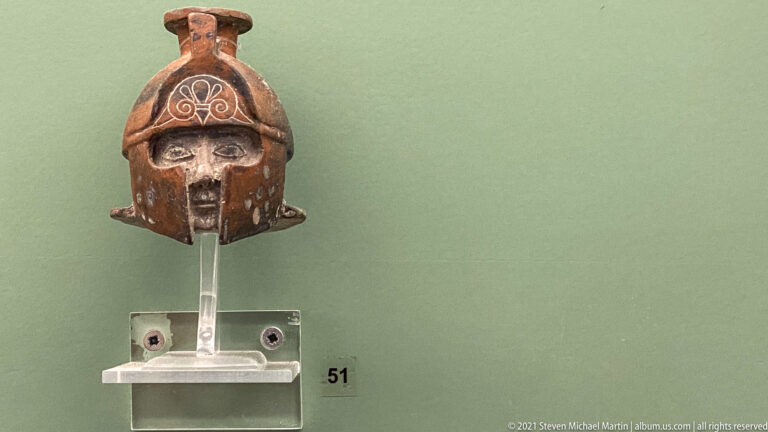 Small dark ceramic helmet-shaped object with frontal opening, side projections, and white painted scroll decoration.
