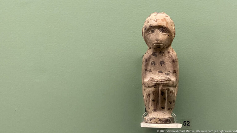 Small upright human figurine with large eyes, arms held across the torso, and dark dotted body decoration.