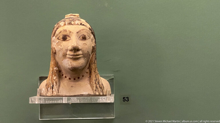 Terracotta female head with long hair, painted eyes and brows, smiling mouth, and dotted neck ornament.