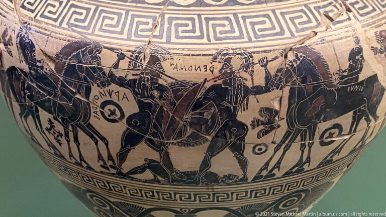Black-figure vase detail with horses, chariots, armed figures, short inscriptions, and meander borders.