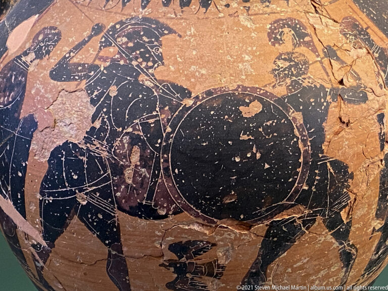 Fragmentary black-figure pottery scene with a large dark animal, partial human figures, abrasion, and missing painted surface.