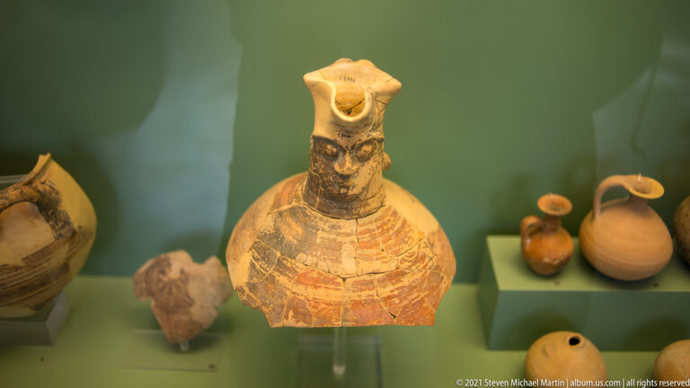 Painted ceramic vessel with modeled human face, rounded body, flaring top, loop handle, and geometric decoration.