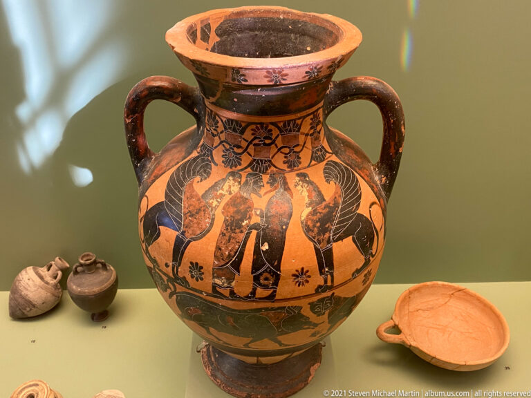 Two-handled black-figure amphora with paired winged figures, floral motifs, dark handles, neck, and foot.