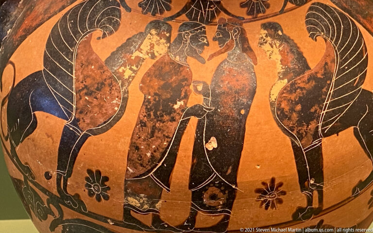 Black-figure pottery detail with standing human figures beneath large birds on an orange clay ground.