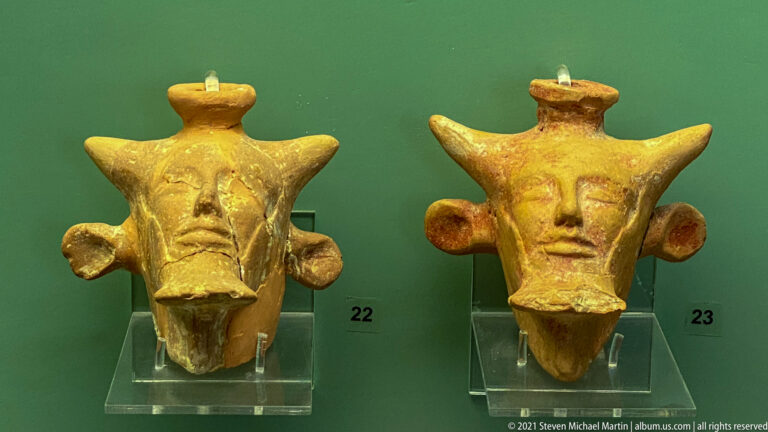 Two small yellow-brown ceramic animal heads with projecting ears or horns, broad muzzles, and top knobs.