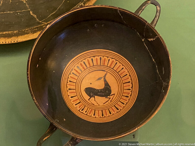Shallow black-glazed handled cup with a central orange medallion containing a dark animal silhouette.