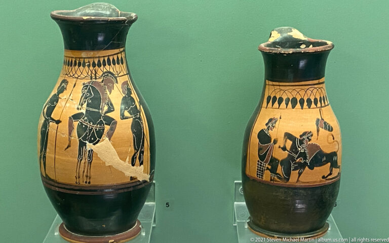Two tall black-figure jugs with dark necks and feet and painted human and animal scenes.