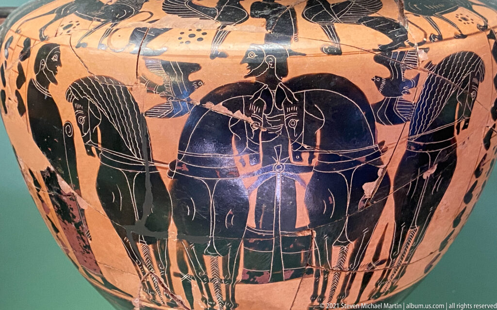Black-figure vase detail with paired horses, chariot figures, and standing attendants on an orange clay ground.