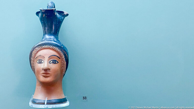 Head-shaped ceramic vessel with modeled female face, painted eyes and lips, dotted hairline, and tall dark neck.