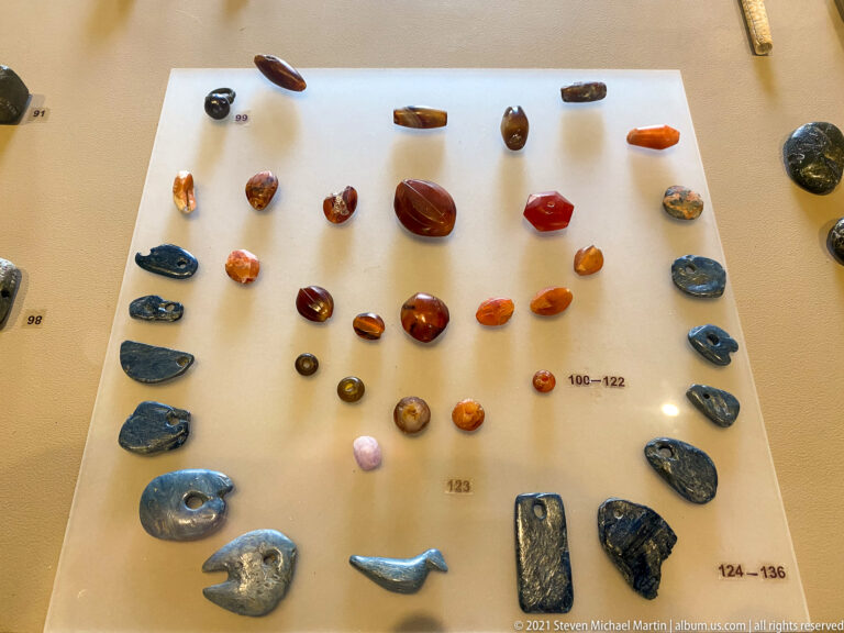 Rows of multicolored beads, carved pendants, polished stones, and small amulets arranged in a museum case.