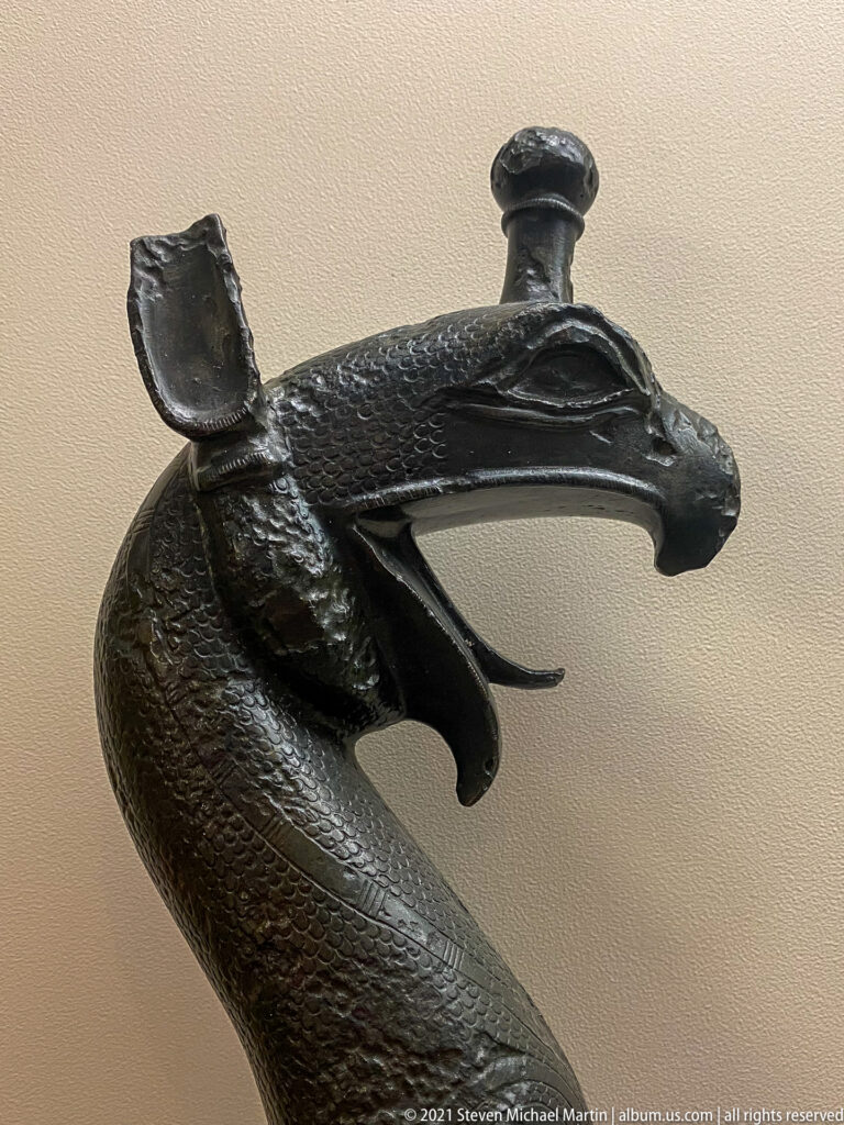 Dark bronze serpentine object ending in an animal head, with projecting fittings and textured surface decoration.