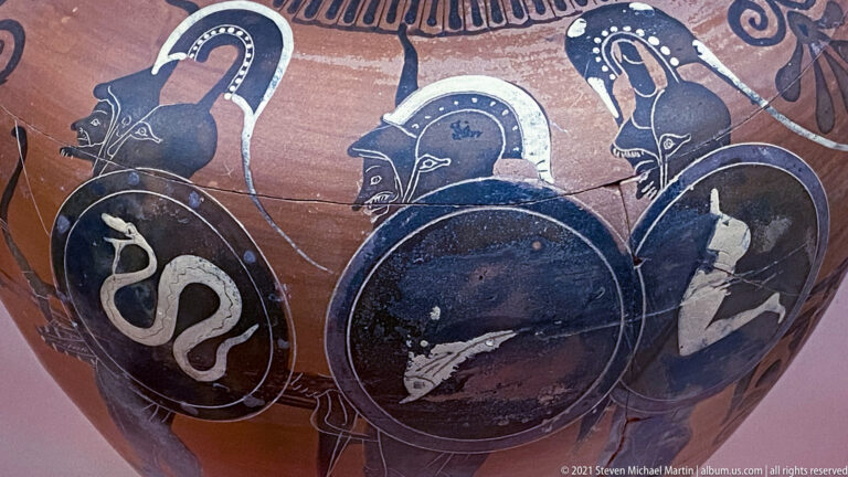 Black-figure warriors carrying round shields with different emblems, including a serpent-like device.