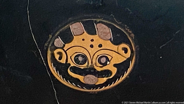 Circular painted medallion with a stylized frontal face, large eyes, broad mouth, and crown-like projections.