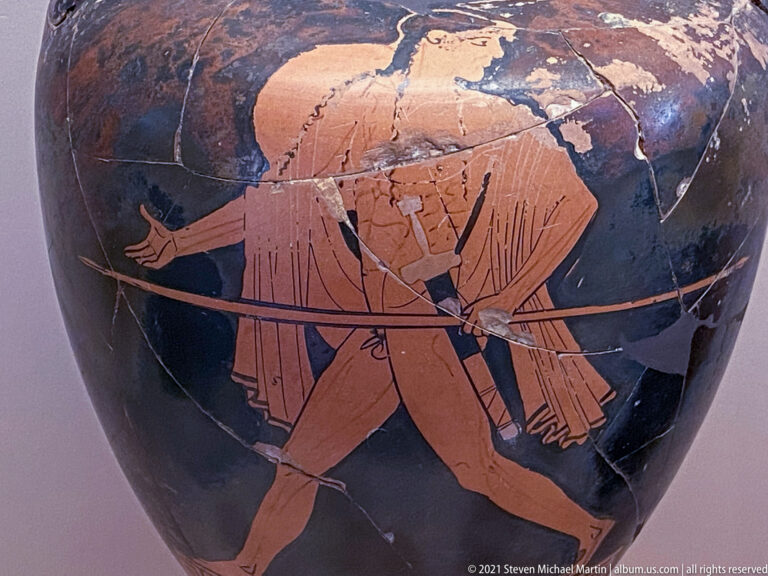 Red-figure warrior striding with a round shield and long spear across a repaired dark vessel surface.