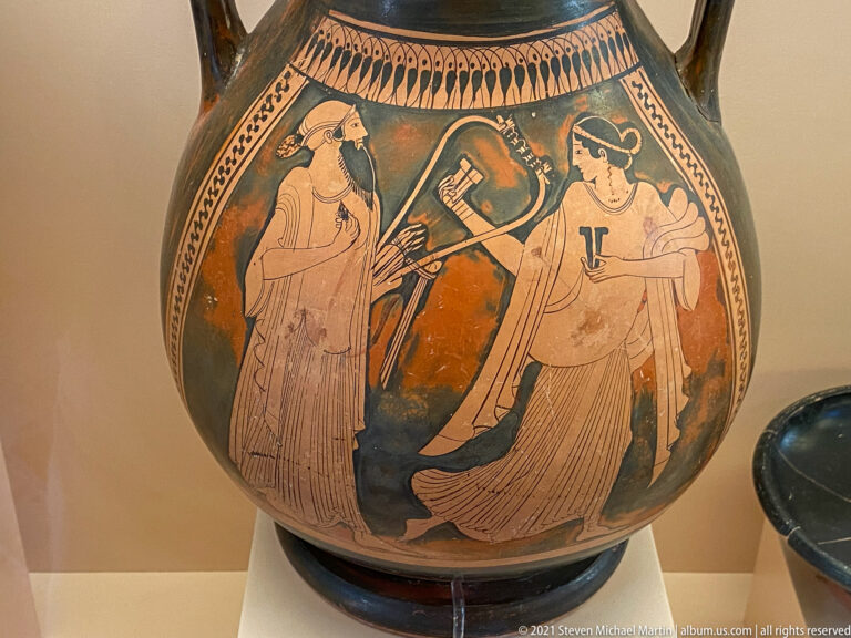 Red-figure vase scene with three draped women grouped around a central object beneath a decorative border.