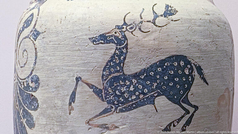 Dark spotted stag in profile with branching antlers and extended legs painted on a pale ceramic surface.
