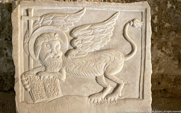 Stone relief of a winged lion-bodied hybrid with a bearded human head beside a small inscribed tablet.